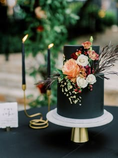 a black and gold wedding cake with flowers on it sitting on a table next to candles