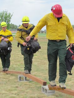 three men in yellow jackets and hard hats are walking across a wooden beam on the grass