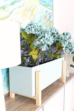 a white planter filled with blue flowers on top of a wooden shelf next to a painting