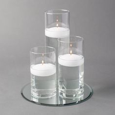 three clear glass vases with white candles in them on a circular plate, set against a gray background