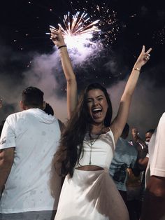 a woman with her arms in the air as fireworks go off behind her and people standing around her