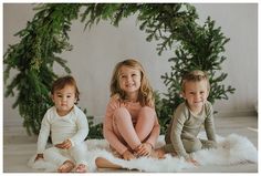 three children are sitting on the floor in front of christmas decorations and greenery, smiling at the camera