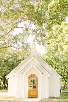 a small white church with a wooden door and window on the front is surrounded by trees