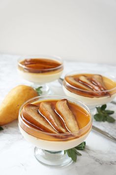 three glasses filled with caramel pudding on top of a marble table next to two pears