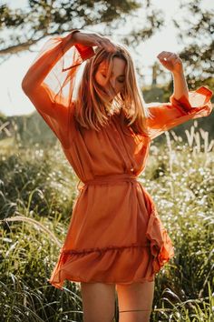 a woman in an orange dress standing in tall grass with her hands behind her head