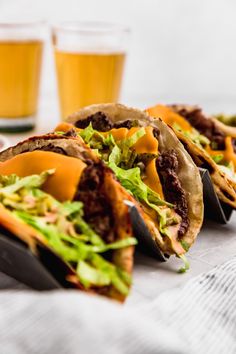 several tacos with lettuce and other toppings sit on a table next to two glasses of beer