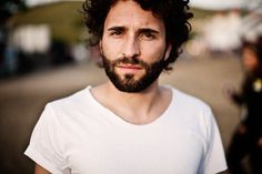 a man with curly hair and beard wearing a white t - shirt looking at the camera