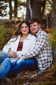 a man and woman sitting on the ground in front of trees with their arms around each other