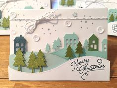 two christmas cards sitting on top of a table next to each other with trees and houses in the background