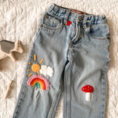 a pair of blue jeans with embroidered rainbows and mushrooms on them, sitting on a bed