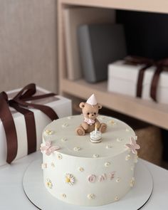 a white cake with a teddy bear on top and a gift box in the background