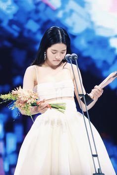 a woman in a white dress is holding a microphone and looking down at her bouquet