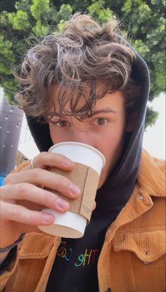 a young man in a hoodie drinking from a paper cup while wearing a brown jacket