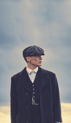 a man wearing a black coat and hat standing in front of a field with tall grass