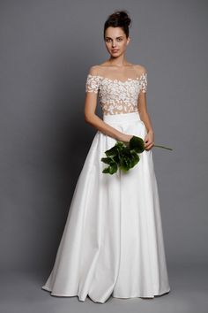 a woman in a white wedding dress holding a flower and looking at the camera while standing against a gray background