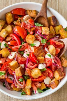 a salad with tomatoes, onions and croutons in a white bowl next to a wooden spoon