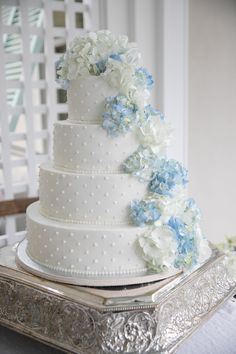 a white wedding cake with blue and white flowers on the top tier is sitting on a silver platter