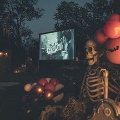 a skeleton sitting in front of a movie screen with balloons and balls on the ground