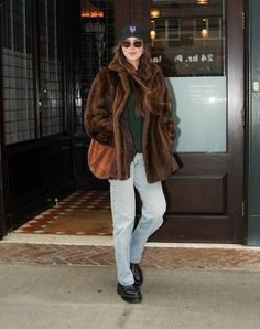 a woman standing in front of a building wearing a fur coat and hat with her hands in her pockets