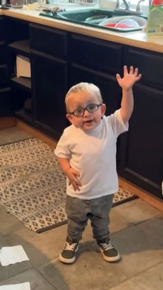 a little boy wearing glasses standing in front of a kitchen counter with his hand up