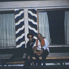 two people are sitting on a bench in front of a building with windows and curtains