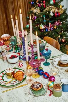 a table is set for christmas dinner with candles on it and other holiday foods in front of the tree