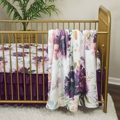 a gold crib with purple and white floral bedding on it, next to a potted plant