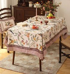 a dining room table set with fruit and flowers on the tablecloth, next to two chairs