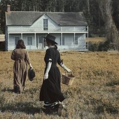 two women walking through a field with a house in the background and one holding a basket