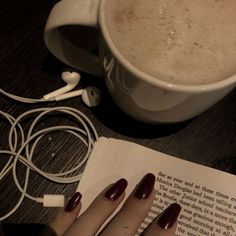 a woman's hands with red nail polish are next to an open book and headphones