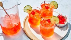 three glasses filled with drinks on top of a white plate next to a bowl of cherries