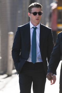 a man in a suit and tie walking down the street with another man wearing sunglasses