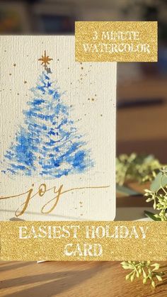 a blue christmas tree on a white card next to some greenery and gold glitter