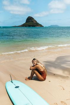 a woman sitting on the beach next to a surfboard with an island in the background