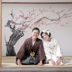 two people sitting on the ground in front of a tree with pink flowers and a painting behind them