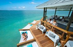 two people are sitting on the deck of a house overlooking the ocean with lounge chairs