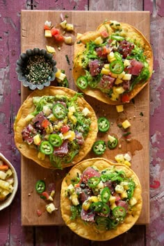 three tacos on a wooden cutting board next to a bowl of fruit and vegetables