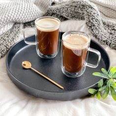 two mugs of coffee sitting on top of a black tray next to a plant