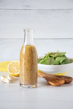 a glass bottle filled with dressing next to a bowl of salad and a wooden spoon