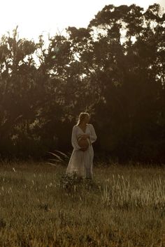 a woman in a white dress holding a basket walking through the grass with trees in the background