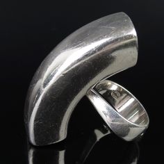 Astrid Fog (b. 1911-d. 1993) designed this 1970s Modernist sleek sculptural sterling ring for Georg Jensen, one of Denmark's most famous silversmith design companies. It has an large curvy elongated futuristic shape that is very shiny and reflective. The back has a curvy sturdy band mounted to it. The inside is signed "Georg Jensen" (incused lettering inside of a dotted oval - hallmark attributed to Georg Jensen beginning 1945). In my photo there was a little residue smudge over the hallmark, wh Formal Sculptural Silver Jewelry, Modernist Silver Dome Ring Open Style, Modernist Sterling Silver Rings For Formal Occasions, Modern Open Bypass Ring For Formal Events, Modern Open Bypass Ring For Formal Occasions, Modern Bypass Open Ring For Formal Occasions, Modern Bypass Ring For Anniversary, Modernist Sterling Silver Rings, Modern Silver Dome Ring