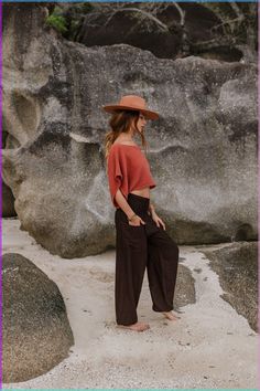 a woman standing in front of large rocks wearing a brown hat and wide legged pants