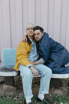 two men sitting on a bench in front of a pink wall and one is hugging the other