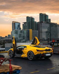 a yellow sports car with its doors open in front of a cityscape at sunset