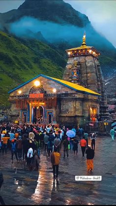 many people are walking around in front of a small temple with mountains in the background