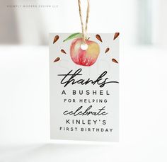 a thank card hanging from a string with an apple on it's tag that says, thanks a bushel for helping celebrate kinley's first birthday