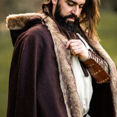 a man with long hair and beard wearing a brown cape is standing in the grass