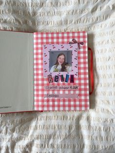 an open book with a photo inside on a white bed sheet next to a red and white checkered blanket