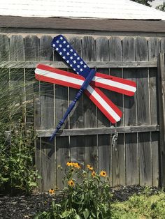 an american flag kite hanging on the side of a wooden fence in front of flowers