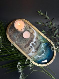 two candles sitting on top of a tray next to some flowers and plants with leaves